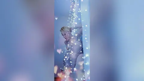 AvaLindt poses naked behind a sheer white curtain decorated with heart-shaped lights, running her hand through her blonde hair.
