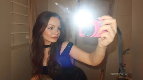 Young woman with long brown hair in a blue tank top taking a selfie in a bathroom mirror.