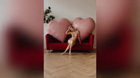 Glamorous woman in pink lingerie relaxing on a red velvet sofa against large heart-shaped pillows.