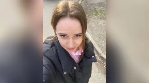 Smiling young woman in a black puffer jacket and pink shirt standing outdoors on a casual day.