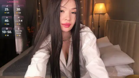 Young woman with long dark hair wearing a white shirt in a cozy, dimly lit bedroom setting.