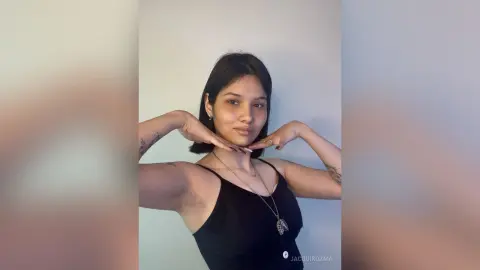 JacquiKozma poses against a white wall, wearing a black tank top and a silver necklace while lifting her arms to show her armpits.