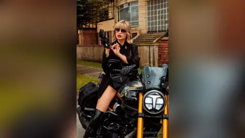 Stylish woman in black outfit and sunglasses posing confidently on a black motorcycle in an urban setting.