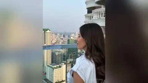 AnaMathers looks out over a city skyline while wearing a white sleeveless top.