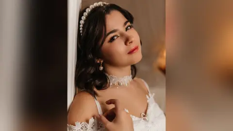 Elegant woman in white lace outfit and pearl headband posing with a soft, romantic glow in a warm indoor setting.