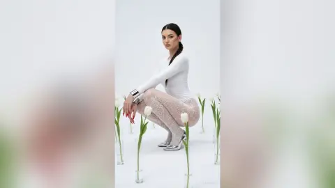 ZenyaSin poses squatting in a white long-sleeve top and patterned tights, surrounded by white tulips on a white background.