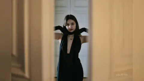 Elegant woman in a black evening dress and scarf posing in a moody, minimalist indoor setting.