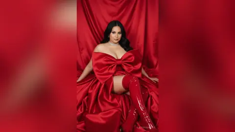Glamorous woman in a red satin gown with a large bow and matching thigh-high boots against a red backdrop.