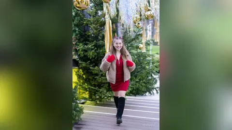 Ada — Smiling woman in a red dress and tan fuzzy coat posing under festive gold Christmas decorations outdoors.
