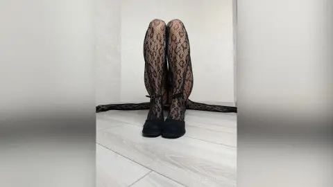 Woman in black lace patterned tights and black heels sitting on a white floor in a minimalist setting.