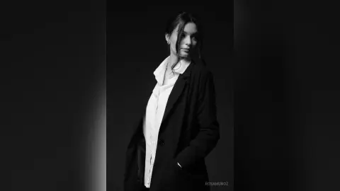 Elegant woman in a black blazer and white shirt posing in a moody black and white studio portrait.