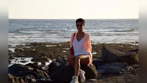 Young woman in a white shirt and pink cardigan sitting on coastal rocks by the ocean at sunset.