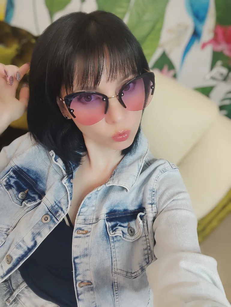 BleckMamba poses with a smirk, wearing black glasses and a black zip-up top with her dark hair styled in a fringe.