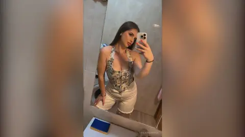 Young woman in a floral corset top and denim shorts taking a mirror selfie in a modern bathroom.