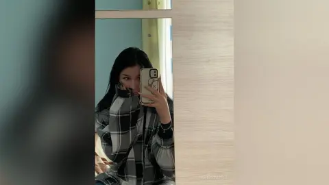 Young woman in a black and white plaid flannel shirt taking a moody mirror selfie in a modern room.