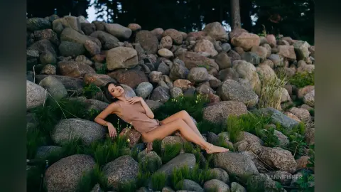 Elegant woman in a beige bodysuit relaxing on a rocky boulder landscape during a serene golden hour sunset.