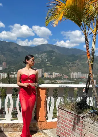 Woman in a red evening dress posing on a sunny balcony overlooking a mountain landscape.