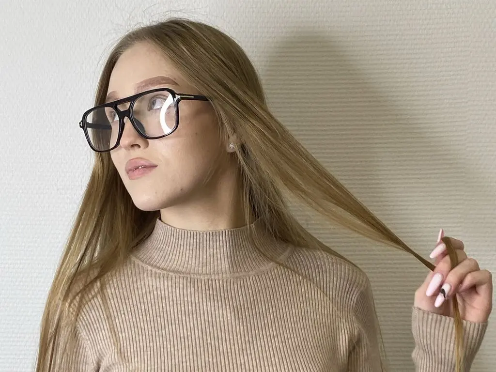 Blonde woman in a beige ribbed turtleneck sweater and black glasses posing against a white wall.