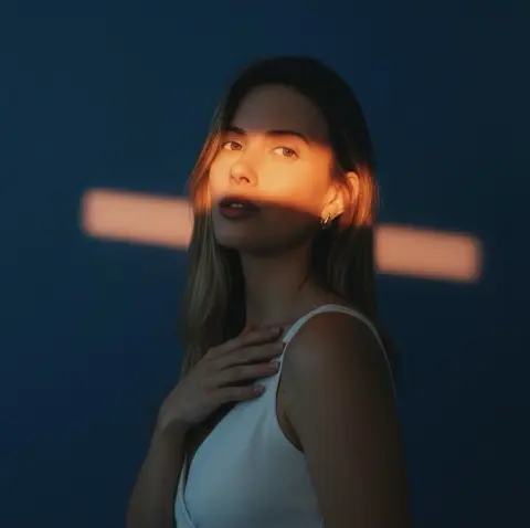 Young woman in a white tank top under dramatic cinematic light against a deep blue background.