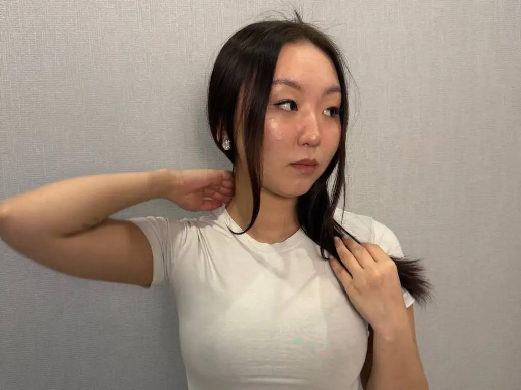 Young woman in a white t-shirt posing against a grey wall with a thoughtful expression.