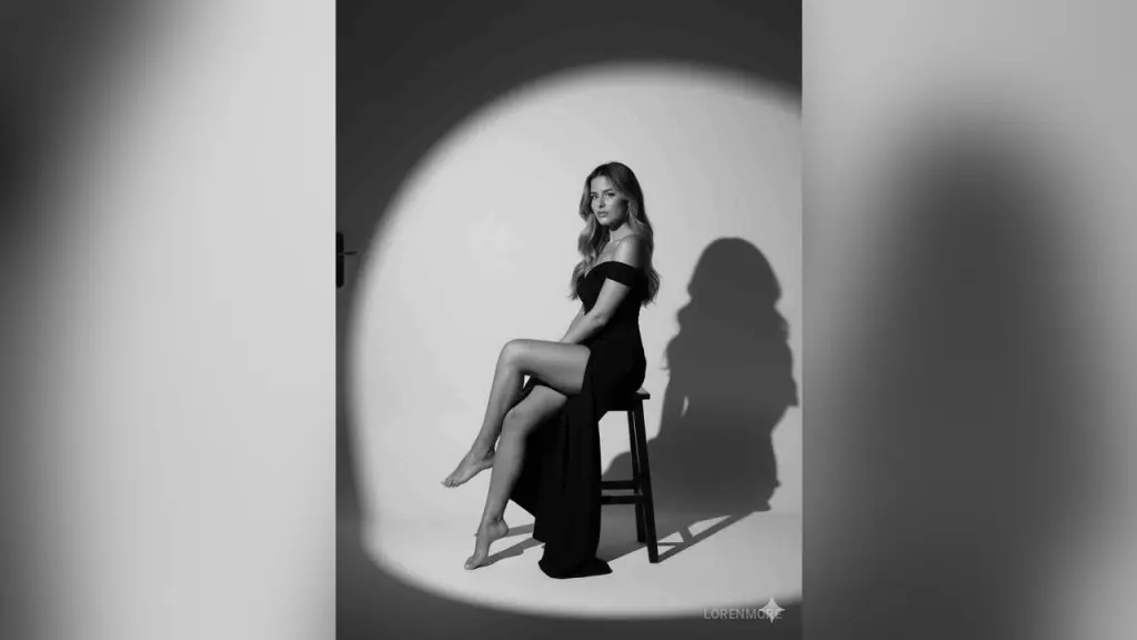 Elegant woman in a shimmering gold cocktail dress relaxing on a black leather chair in moody lighting.