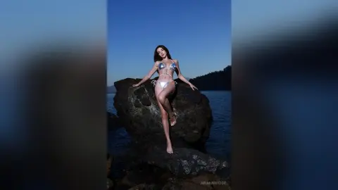 Slim woman in a silver bikini posing on dark rocks by the ocean under a twilight blue sky.