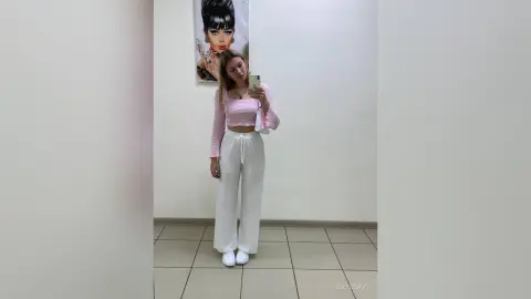 Young woman in a pink long-sleeve crop top and white wide-leg lounge pants taking a mirror selfie in a bright room.