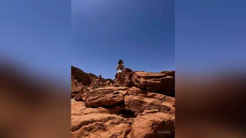 Young woman in white summer outfit standing on red desert rocks under a clear blue sky.