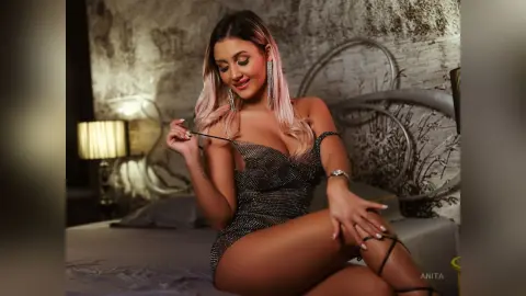 Anita sits on a bed wearing a black mesh dress with silver sparkles, looking down while touching her strap.
