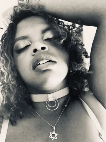 Close-up portrait of a woman with curly hair wearing a choker and Star of David necklace in a moody black and white style.