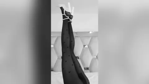 Woman in black tights and white high heels posing with legs raised in a bright, modern bedroom setting.
