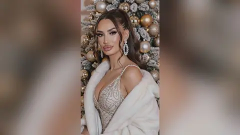 AnnaKarev poses in a sparkly cleavage-baring dress and white fur coat in front of a decorated Christmas tree.