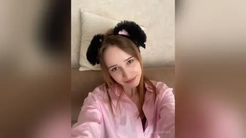 Young woman in a pink button-down shirt wearing black fuzzy cat ears in a cozy bedroom setting.