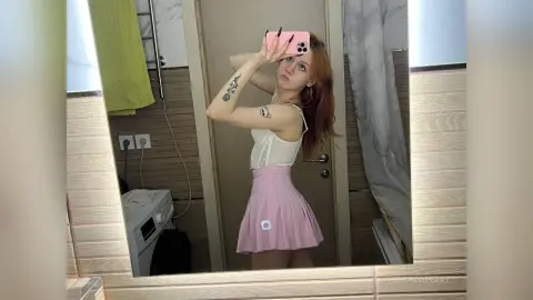 Young woman in a white tank top and pink pleated skirt taking a mirror selfie in a modern bathroom.