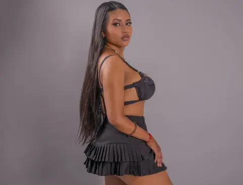 Young woman with long dark hair wearing a black crop top and pleated mini skirt in a studio setting.
