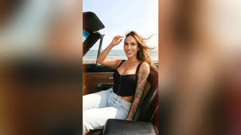 LonisMillers smiles while sitting in a car, wearing a black tank top and light blue jeans with her long hair blowing in the wind.