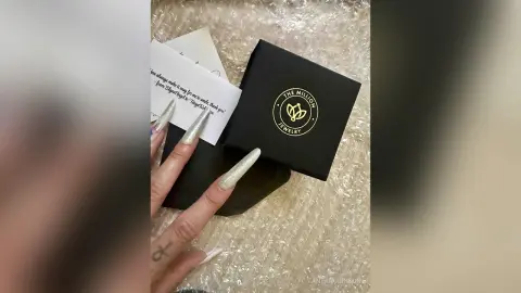 Hand with long silver glitter nails opening a black The Million Jewelry gift box on white bubble wrap.
