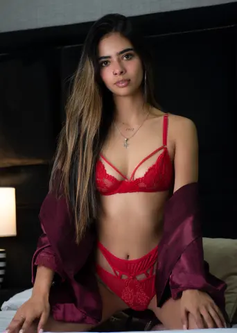 Young woman in red lace lingerie and a maroon silk robe sitting on a bed in a moody bedroom setting.