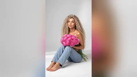 Blonde woman in denim jeans holding a large bouquet of pink roses against a white studio background.