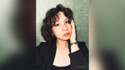 Young woman with wavy brown hair wearing a black blazer posing against a textured green wall with a soft mood.