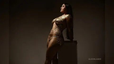 Elegant woman in a gold sequin bodysuit posing in a moody, dark studio setting.