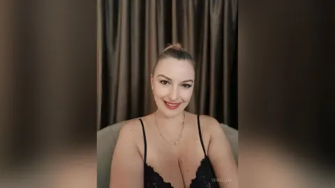 VeraLuxe — VeraLuxe smiles at the camera wearing a black lace camisole and a gold necklace against dark curtains.