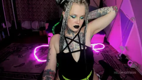 Tattooed woman with silver braids in a black bodysuit posing in a moody, neon pink lit room.
