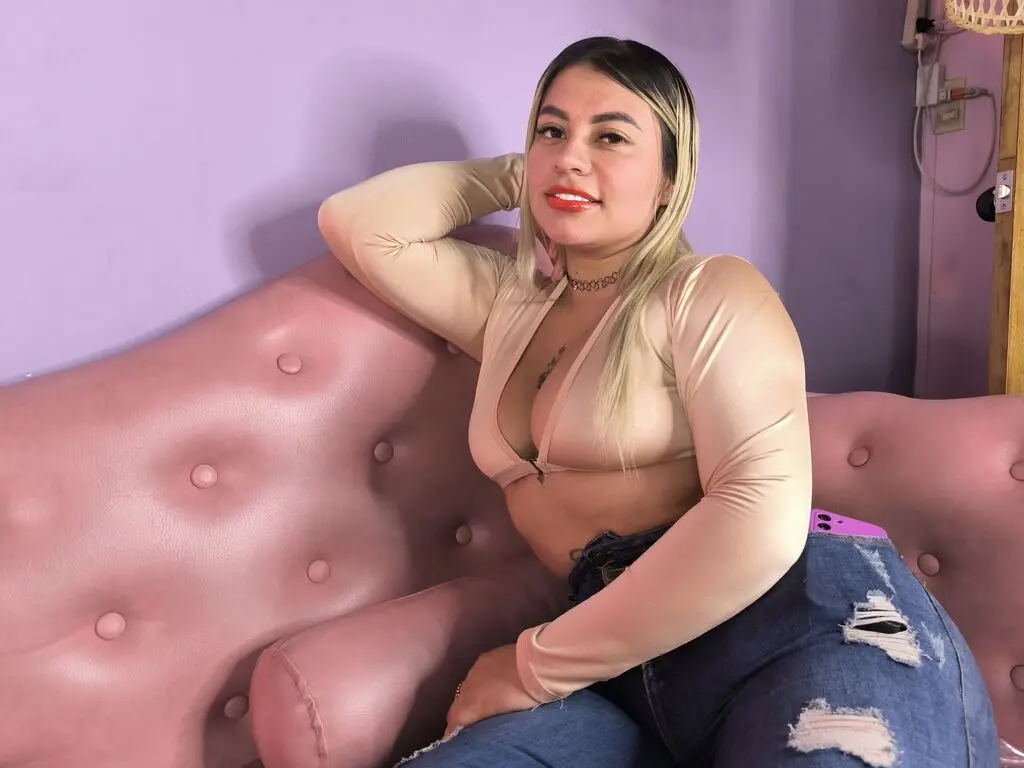 Smiling woman in a beige long-sleeve crop top and ripped blue jeans sitting on a pink velvet sofa.