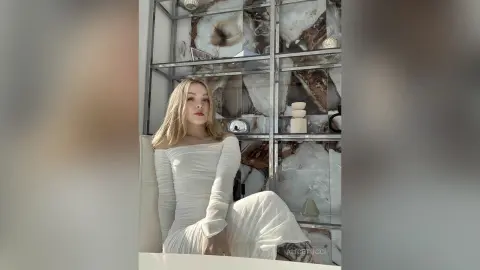AliceTucci poses in a white long-sleeved dress with her blonde hair down, sitting in front of a decorative glass display shelf.