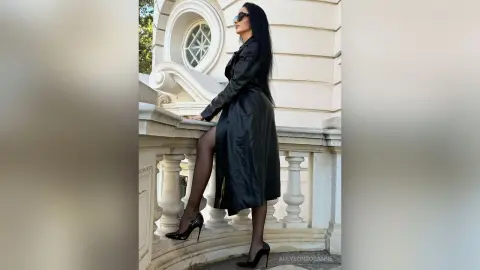 Elegant woman in a long black leather trench coat and heels posing by a classic white stone balcony.
