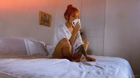 Redheaded woman in white shirt sipping coffee on a bed in a cozy, sunlit bedroom.
