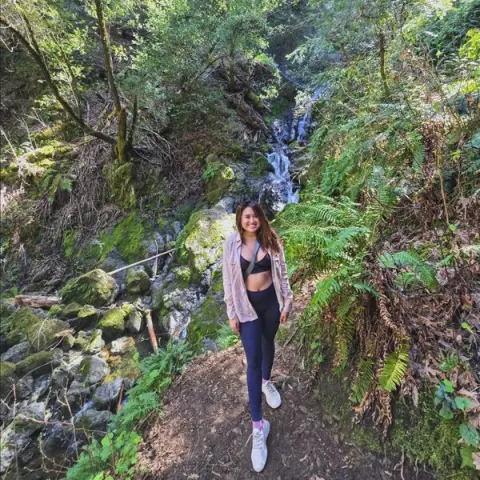 Smiling woman in athletic wear hiking on a lush forest trail near a scenic mountain waterfall.