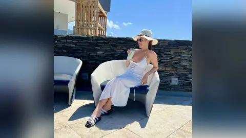 Stylish woman in a white summer dress and sun hat relaxing on a sunny rooftop patio with a refreshing drink.