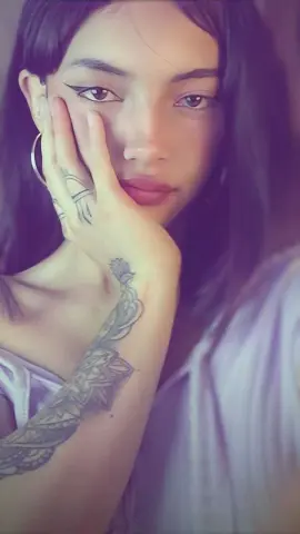 Young woman with long dark hair and arm tattoos posing in a white top with a soft, moody aesthetic.